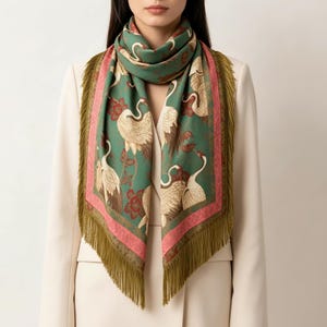 May include: A square scarf featuring a green background with a pattern of white cranes and red floral accents. The scarf is bordered by a pink band and finished with long olive green fringe along the edges, draped around the neck.
