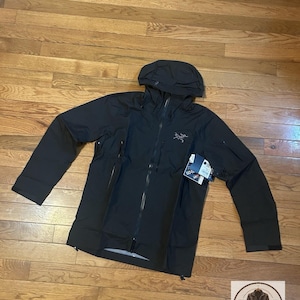 Puede incluir: Una chaqueta negra con capucha que presenta una cremallera frontal completa, mangas largas con puños ajustables y un logotipo en el pecho. La prenda incluye una etiqueta Gore-Tex adjunta y varios bolsillos con cremallera en los laterales.