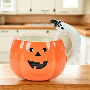 Peut inclure: Un mug en céramique en forme de citrouille orange avec un visage de jack-o'-lantern aux yeux noirs, un nez triangulaire et une bouche dentelée. L'anse est conçue comme un fantôme blanc portant un petit chapeau noir, incurvé sur le côté du mug.