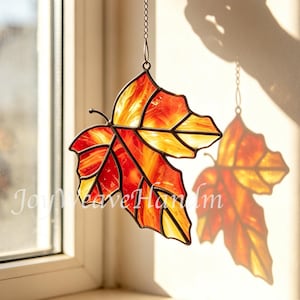 Op de afbeelding: Een glas-in-lood zonnevanger in de vorm van een esdoornblad met levendige rode, oranje en gele glassegmenten die bij elkaar worden gehouden door donkere metalen soldeerlijnen. Het blad hangt aan een dunne metalen ketting.
