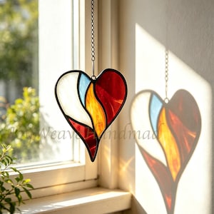 May include: A heart-shaped stained glass suncatcher featuring segments of white, light blue, yellow, and red glass held together by dark metal cames. The piece hangs from a thin metal chain and casts a colorful shadow on the adjacent wall.