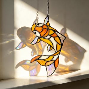 May include: A stained glass suncatcher shaped like a koi fish, featuring vibrant orange, yellow, and iridescent white glass segments held together by dark metal solder lines, suspended by a thin chain to catch and refract natural sunlight.
