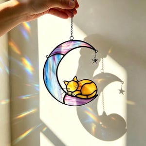 Könnte beinhalten: Ein Buntglas-Sonnenfänger in Form eines Halbmondes in Violett-, Blau- und Weißtönen mit einer schlafenden gelben Katze auf der Kurve. Ein kleiner Metallstern hängt am Mond, und das gesamte Stück ist an einer Metallkette befestigt.