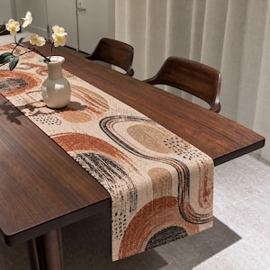 Op de afbeelding: Een rechthoekige tafelloper met een beige basis en een herhalend abstract patroon van gebogen lijnen en cirkelvormige figuren in tinten van gebrand oranje, terracotta en zwart, liggend op een donkerhouten eettafel.