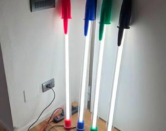 Peut inclure: Quatre lampes sur pied en forme de stylos à bille géants. Chaque lampe présente un corps cylindrique transparent avec une lumière blanche vive, surmonté d'un capuchon rouge, bleu, vert ou noir, avec des bases de couleurs assorties.