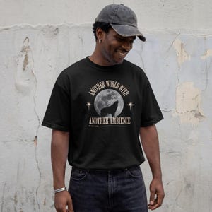 Pode incluir: Uma t-shirt preta de manga curta com uma estampa gráfica de um lobo a uivar para a lua cheia. O texto "ANOTHER WORLD WITH ANOTHER AMBIENCE" está impresso numa fonte serifada bege acima e abaixo da lua, com pequenos ícones de estrelas.