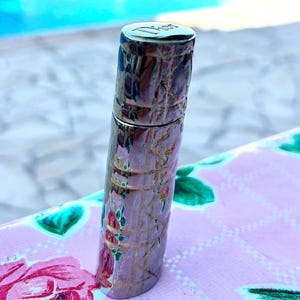 Pode incluir: Um estojo de batom Dior cilíndrico com acabamento prateado metálico, apresentando um padrão cannage em relevo e um delicado design floral de rosas cor-de-rosa e folhas verdes. O nome da marca Dior está gravado no topo da tampa.