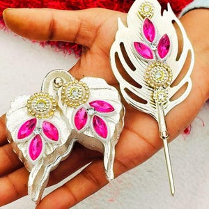 Puede incluir: Un conjunto de accesorios para el cabello de metal plateado que incluye un alfiler con forma de hoja y una pieza decorativa a juego de dos hojas. Ambos artículos están adornados con cristales rosas y brillantes transparentes.