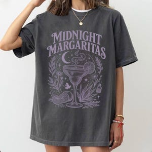Puede incluir: Una camiseta gris carbón de manga corta con un estampado gráfico en color morado claro. El diseño incluye el texto "MIDNIGHT MARGARITAS" sobre una ilustración de una copa de margarita, una luna creciente, estrellas, flores y cítricos.