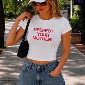 May include: A white short-sleeved t-shirt with the red text "RESPECT YOUR MOTHER!" printed on the chest, paired with a white baseball cap, brown sunglasses, and a black shoulder bag, worn with blue denim jeans.