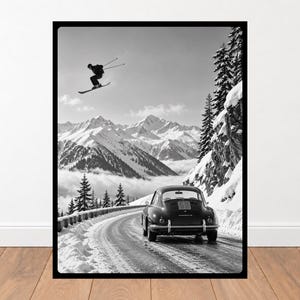 May include: A black and white framed wall art print featuring a vintage sports car driving on a snowy mountain road. Above the car, a skier is captured mid-air while jumping. The background displays snow-covered mountain peaks and pine trees.