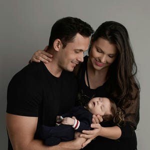 May include: A family of three with two adults wearing black shirts and a sleeping infant dressed in a navy blue onesie with white and red striped cuffs. The infant is held securely in the arms of the adults against a plain gray background.