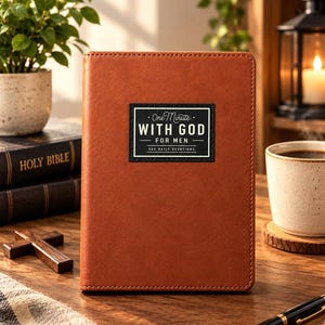 May include: A brown leather-textured devotional book with a rectangular black label on the front cover. The label features the white text "One Minute WITH GOD FOR MEN 365 DAILY DEVOTIONS" with decorative lines and a border centred on the cover.