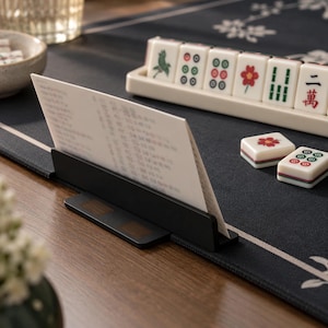 May include: A black metal card holder stand displaying a white card with printed text, positioned on a dark fabric mat alongside a cream-colored tile rack containing several mahjong tiles with green, red, and black symbols and characters.