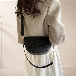 Peut inclure: Un sac bandoulière noir en forme de selle avec une finition lisse et un rabat incurvé. Le sac est doté d'une large bandoulière en tissu noir et d'une seconde chaîne décorative en métal doré fixée sur les côtés du sac.