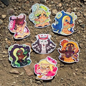 May include: A collection of seven die-cut stickers featuring stylized cartoon characters with vibrant hair colors including pink, green, blue, purple, orange, and yellow. Each character has distinct facial expressions and holographic glitter details.
