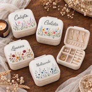 May include: Four square, cream-colored jewelry boxes with floral prints and personalized names including Calista, Linnea, and Melisande. One box is open, revealing a beige interior with hooks for necklaces, small compartments, and ring slots.