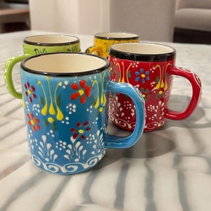 Op de afbeelding: Een set van vier keramische mokken in blauw, rood, geel en groen, elk met een zwarte rand en een handgeschilderd bloemmotief met witte, gele en rode details. De mokken hebben een cilindrische vorm met een gebogen handvat aan de zijkant.