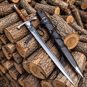 May include: A steel sword with a polished blade, a cross-shaped guard featuring a small shield emblem, and a brown wooden handle with metal accents. Beside the sword lies a black leather scabbard with leather straps and a metal tip.