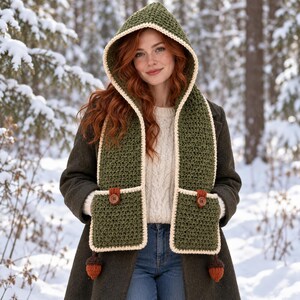 May include: A hooded crochet scarf in olive green with cream-colored trim. The scarf features two front pockets, each secured with a brown button and a small brown tab. Two small, dark brown acorn-shaped tassels hang from the bottom of each pocket.