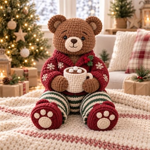 Könnte beinhalten: Ein brauner gehäkelter Teddybär in einer roten Strickjacke mit weißen Schneeflockenmustern und einer grün-weiß gestreiften Hose. Der Bär hält eine cremefarbene Tasse mit einem braunen Getränk, das mit drei weißen Marshmallows garniert ist.