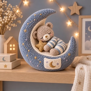Könnte beinhalten: Ein gehäkelter Halbmond in Blau mit cremefarbenem Innenbereich und gestickten goldenen Sternen. Ein kleiner, hellbrauner gehäkelter Teddybär mit geschlossenen Augen und blau-weiß gestreiftem Schlafanzug liegt darin und hält einen goldenen Stern.