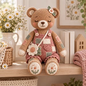 Può includere: Un orsacchiotto all'uncinetto in filato marrone, che indossa una salopette rosa con quadrati patchwork e un dettaglio a cuore. L'orso ha un fiore di margherita sulla testa e porta una borsa a tracolla abbinata. I piedi sono decorati con motivi a margherita ricamati.