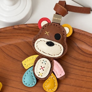 May include: A brown leather bear keychain featuring a patchwork design with yellow, blue, and pink limbs. The bear has a cream-colored muzzle, a black nose, one black eye, and a yellow stitched X over the other eye. It includes a brown leather strap.