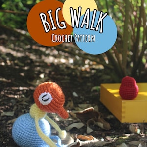 Peut inclure: Un modèle de crochet intitulé BIG WALK représentant une petite créature faite à la main, multicolore, avec une tête orange ronde, un corps bleu clair et des membres jaunes fins. Un petit objet rouge au crochet repose sur une boîte jaune.
