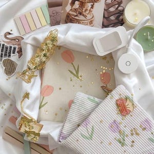 Puede incluir: Una colección de artículos de papelería que incluye un cuaderno color crema con ilustraciones de tulipanes rosas, un estuche blanco acanalado con patrones de tulipanes morados, confeti de estrellas doradas, pines decorativos, notas adhesivas y dos velas.