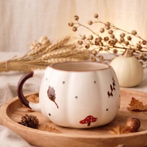 Puede incluir: Una taza de cerámica color crema con forma de calabaza y asa marrón oscuro. La taza presenta un patrón de pequeños puntos grises, un hongo rojo con puntos blancos e ilustraciones de hojas marrones pintadas en la superficie exterior.