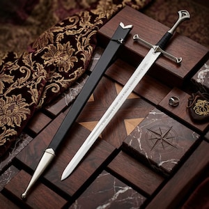May include: A silver-toned metal sword with a black grip and a crossguard, accompanied by a black scabbard with silver-toned metal fittings. The blade features engraved text and a small sun symbol near the hilt, alongside a small silver ring.