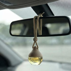 May include: A car air freshener hanging from a rearview mirror, featuring a small, clear glass bottle filled with a golden liquid, topped with a light-colored wooden cap and suspended by a beige cord with an adjustable wooden bead.