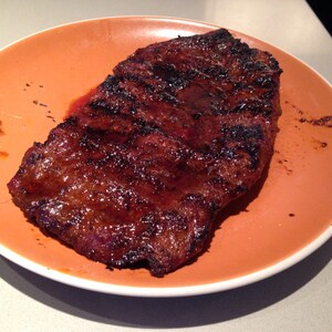 May include: A grilled steak with a dark, charred exterior and visible grill marks, served on a round, solid orange ceramic plate. The meat has a glossy, cooked appearance with deep brown and black tones, resting centrally on the smooth, matte-finish plate.