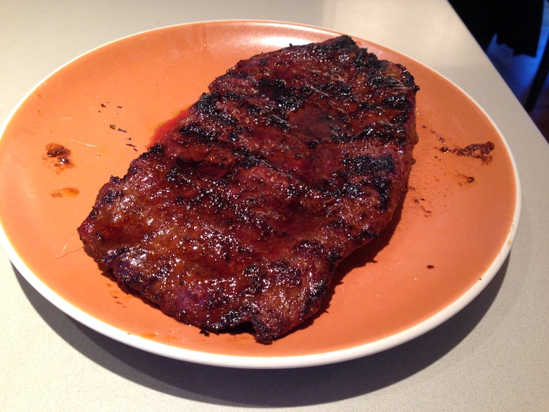 May include: A grilled steak with a dark, charred exterior and visible grill marks, served on a round, solid orange ceramic plate. The meat has a glossy, cooked appearance with deep brown and black tones, resting centrally on the smooth, matte-finish plate.