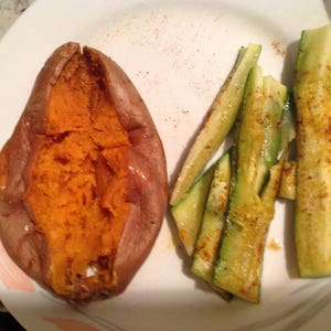 May include: A baked sweet potato with a split, orange interior and brown skin, served alongside several seasoned, sliced green zucchini strips on a white plate with subtle pink and grey decorative accents near the rim.