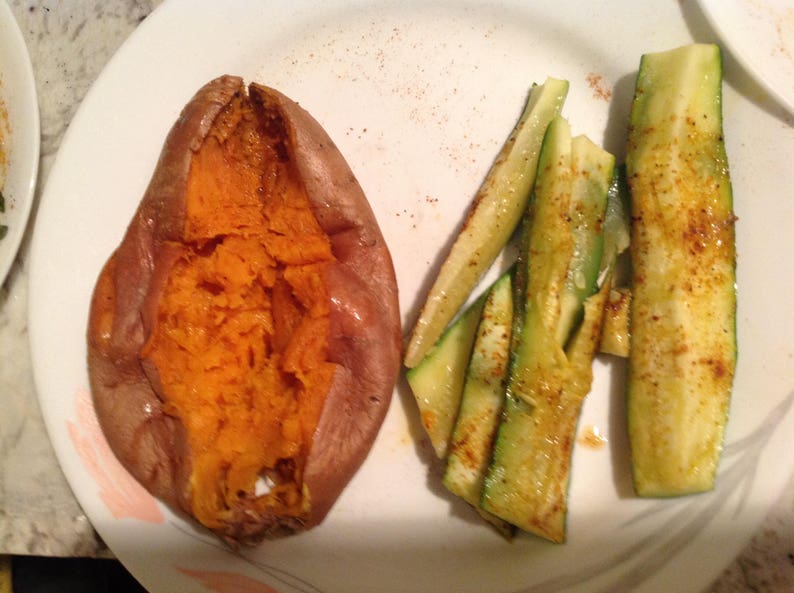 May include: A baked sweet potato with a split, orange interior and brown skin, served alongside several seasoned, sliced green zucchini strips on a white plate with subtle pink and grey decorative accents near the rim.