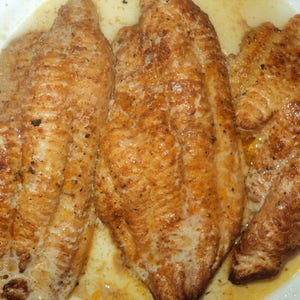 May include: Three golden-brown pan-seared fish fillets with a seasoned, textured surface. The fillets are cooked to a medium-brown color and appear moist, with visible flecks of dark seasoning spices distributed across the surface of the meat.