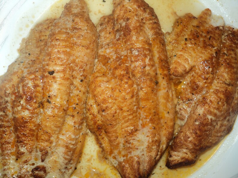 May include: Three golden-brown pan-seared fish fillets with a seasoned, textured surface. The fillets are cooked to a medium-brown color and appear moist, with visible flecks of dark seasoning spices distributed across the surface of the meat.