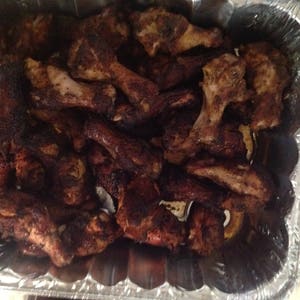 May include: A large quantity of cooked chicken wings with a dark, seasoned, and charred exterior, packed tightly into a rectangular, disposable aluminum foil tray. The wings show varying shades of deep brown and black from the cooking process.