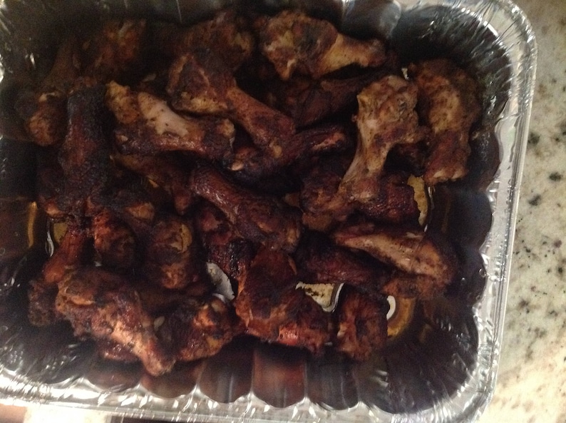 May include: A large quantity of cooked chicken wings with a dark, seasoned, and charred exterior, packed tightly into a rectangular, disposable aluminum foil tray. The wings show varying shades of deep brown and black from the cooking process.