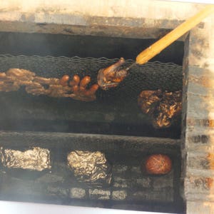 May include: A brick-lined barbecue pit with two metal grates. The top grate holds cooked meat, sausages, and a piece of poultry on a wooden skewer. The bottom grate contains two items wrapped in silver foil and a round, reddish-brown food item.