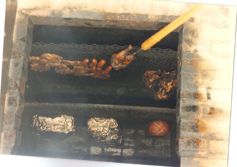 May include: A brick-lined barbecue pit with two metal grates. The top grate holds cooked meat, sausages, and a piece of poultry on a wooden skewer. The bottom grate contains two items wrapped in silver foil and a round, reddish-brown food item.