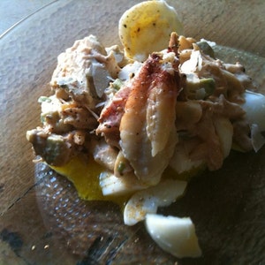 May include: A seafood salad featuring a prominent crab claw, chunks of white fish, and sliced hard-boiled eggs, all coated in a creamy dressing with visible black pepper and thin shavings of white cheese, served on a clear textured glass plate.