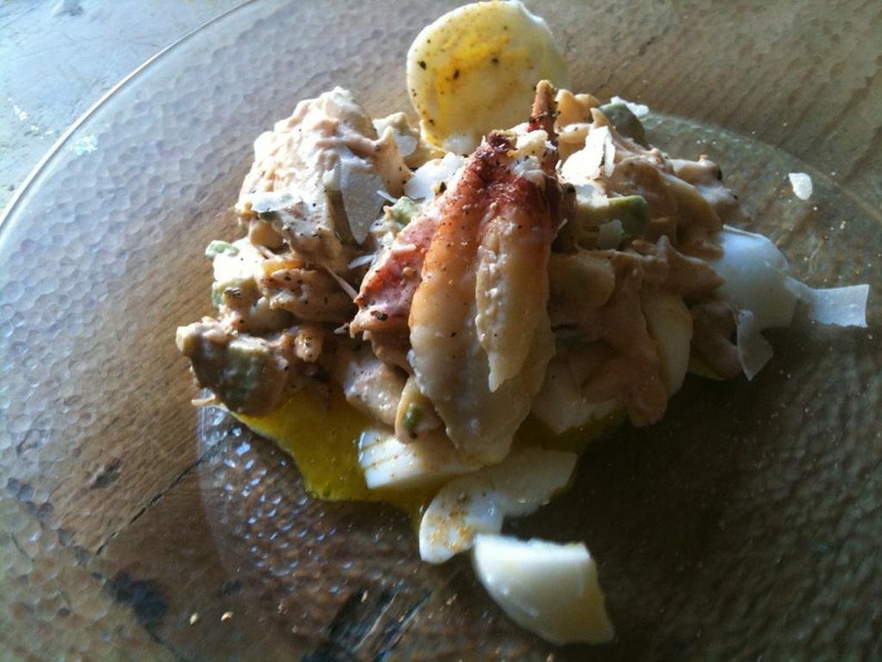 May include: A seafood salad featuring a prominent crab claw, chunks of white fish, and sliced hard-boiled eggs, all coated in a creamy dressing with visible black pepper and thin shavings of white cheese, served on a clear textured glass plate.