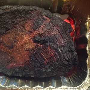 May include: A large, cooked beef brisket with a dark, charred, and textured bark surface, resting inside a rectangular aluminum foil pan. The meat shows deep brown and black coloration from the smoking process, with visible muscle fibers.