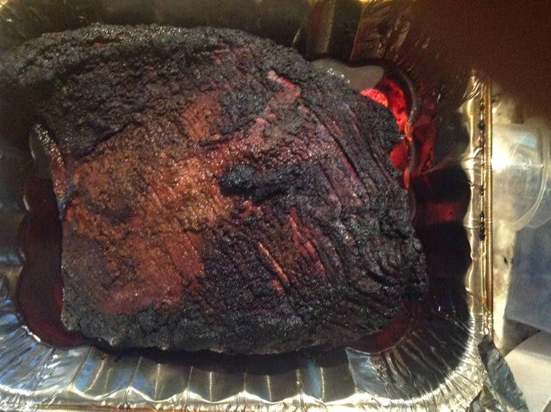 May include: A large, cooked beef brisket with a dark, charred, and textured bark surface, resting inside a rectangular aluminum foil pan. The meat shows deep brown and black coloration from the smoking process, with visible muscle fibers.