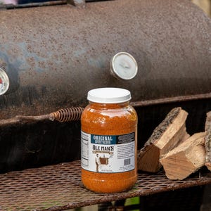 May include: A clear plastic jar filled with orange spice blend labeled Ole Man's Original Spice Blend, positioned on a metal grill grate next to two pieces of firewood, with a rusted metal smoker featuring two circular temperature gauges behind it.