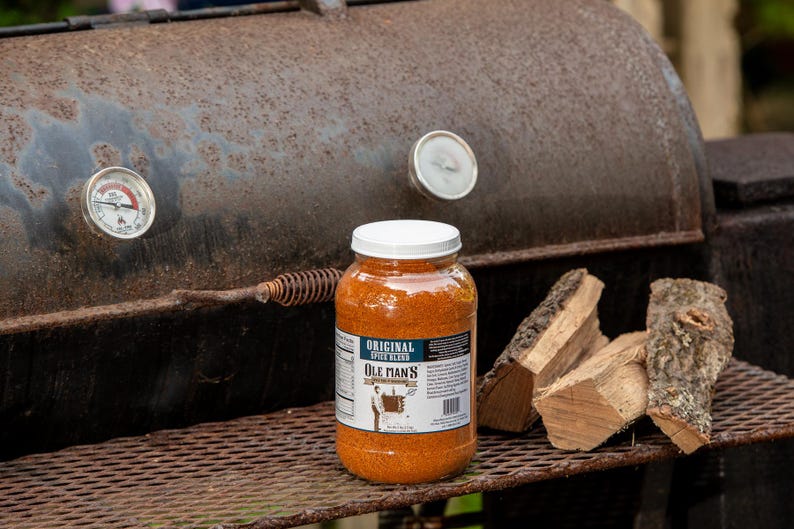 May include: A clear plastic jar filled with orange spice blend labeled Ole Man's Original Spice Blend, positioned on a metal grill grate next to two pieces of firewood, with a rusted metal smoker featuring two circular temperature gauges behind it.