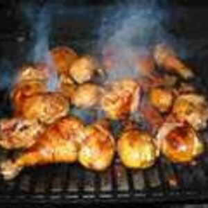 May include: A collection of golden-brown grilled chicken drumsticks and thighs arranged on a black metal barbecue grate. Wisps of white smoke rise from the seasoned poultry as it cooks over the heat, showing charred grill marks on the skin.
